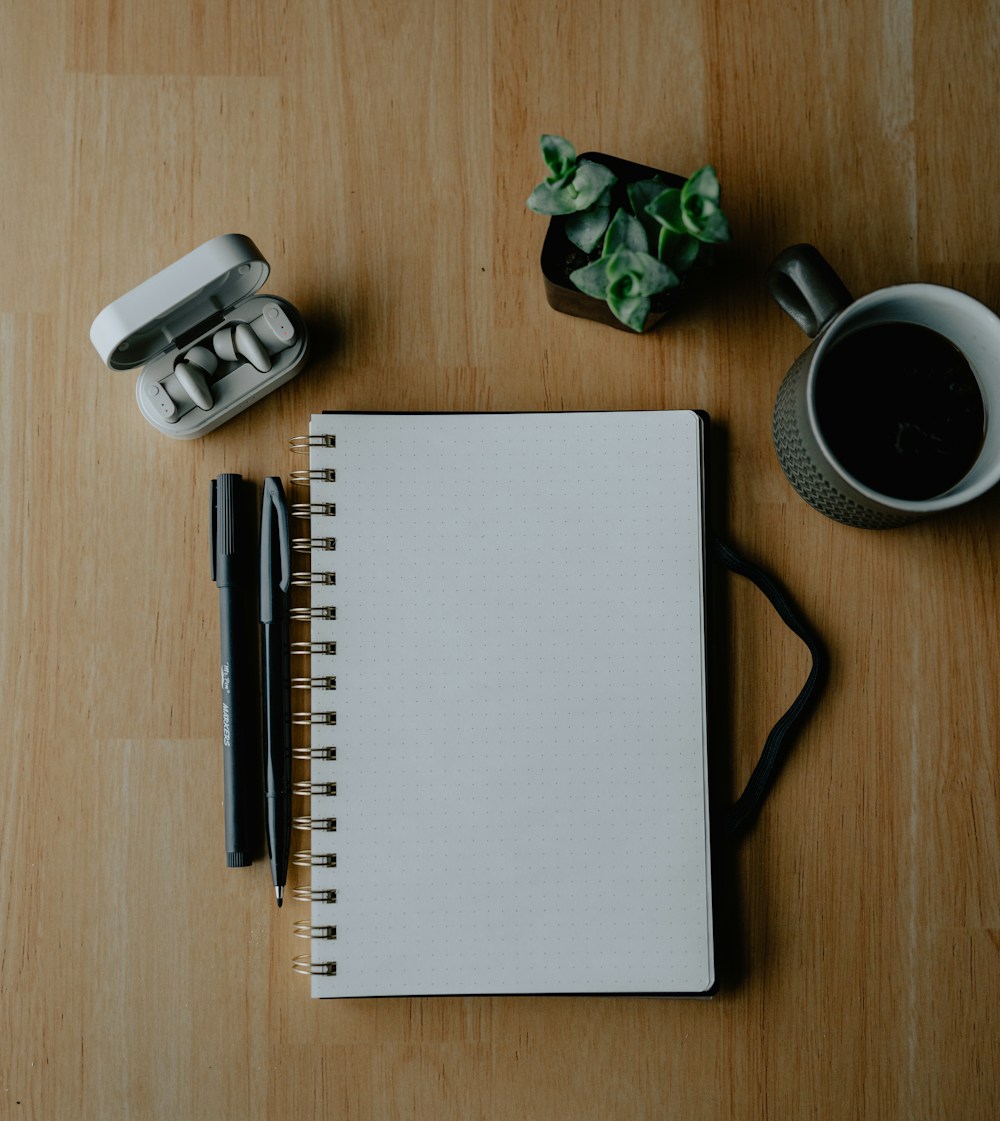 An open notebook, pens and coffee on a wooden desk.